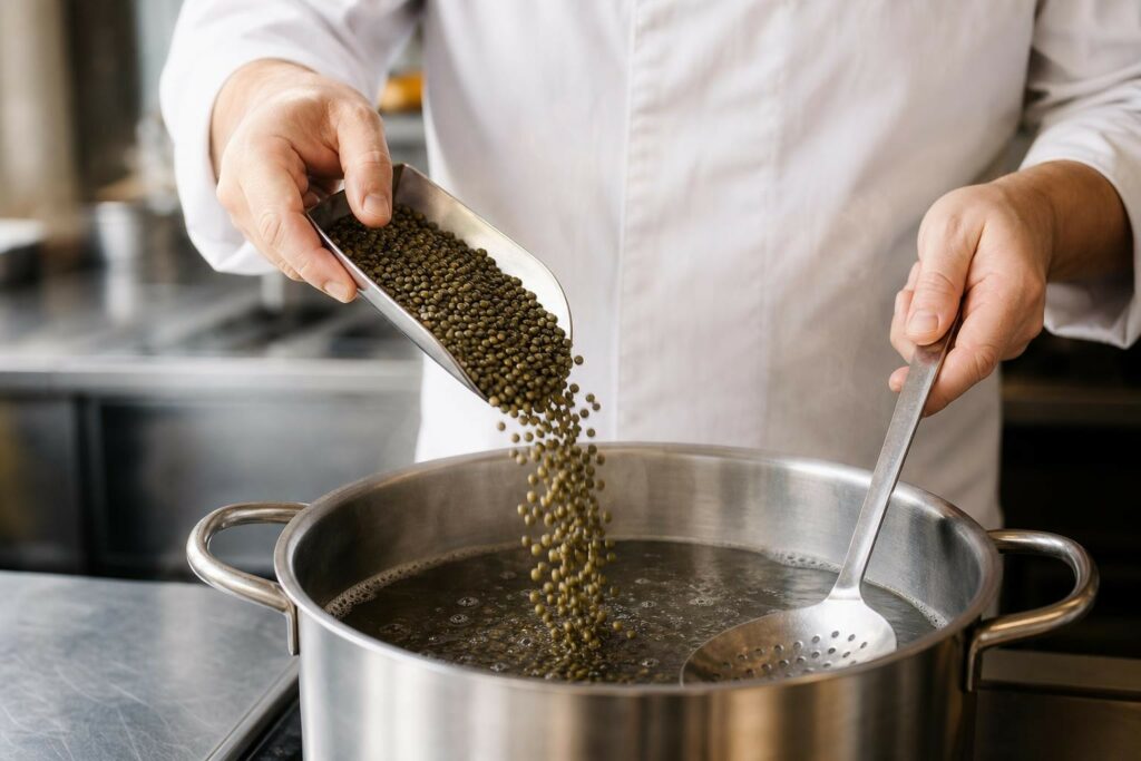 Scène évocatrice de coulisses de cuisine : mains anonymes versant des lentilles vertes du Puy dans un faitout en inox