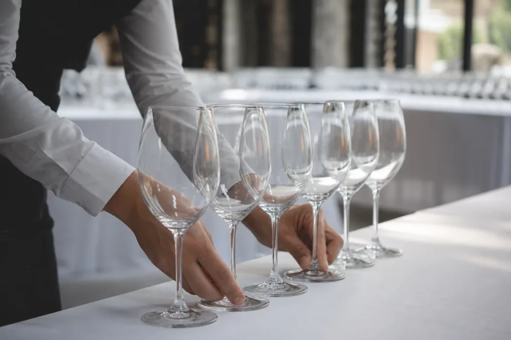 Scène évocatrice des coulisses d’un salon œnologique avec des mains anonymes alignant des verres à vin vides sur une table nappée blanche dans un espace intérieur flou.