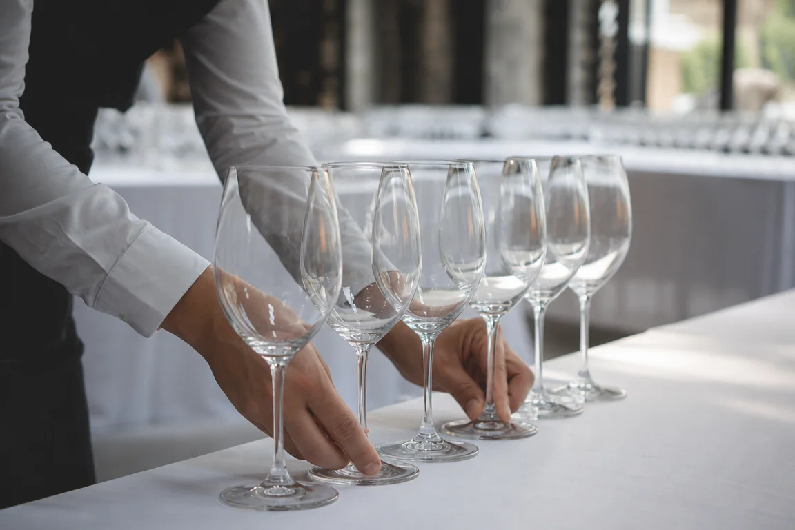 Préparation des verres pour le Salon des Vins de Thoiry Scène évocatrice des coulisses d’un salon œnologique avec des mains anonymes alignant des verres à vin vides sur une table nappée blanche dans un espace intérieur flou.
