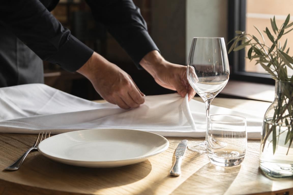 Mains anonymes disposant une nappe blanche et une assiette sur un plan de travail en bois clair, évoquant la préparation en coulisses d’une nouvelle trattoria.
